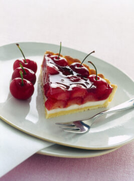 Fetta di crostata con base di frolla, strato di panna cotta bianca e una ricca copertura di ciliegie intere immerse in una gelatina rossa lucida, servita su un piatto bianco con forchettina.