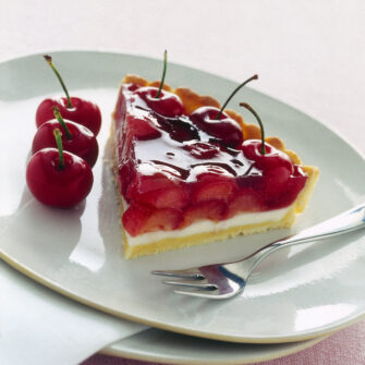 Fetta di crostata con base di frolla, strato di panna cotta bianca e una ricca copertura di ciliegie intere immerse in una gelatina rossa lucida, servita su un piatto bianco con forchettina.