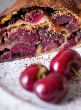 Primo piano di una fetta di strudel ripiena di tantissime ciliegie cotte e scure, con una spolverata di cacao in polvere in superficie e ciliegie fresche in primo piano. 