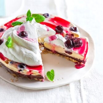 Torta fredda tipo cheesecake alle ciliegie con base scura, crema chiara, abbondante panna montata, topping e ciliegie intere, con una fetta parzialmente tagliata. 