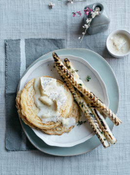 Pancake di avena con sopra camembert fuso, accompagnati da asparagi bianchi grigliati su piatto bianco