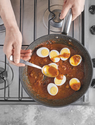 Mano con cucchiaio che versa in padella salsa al curry su uova sode