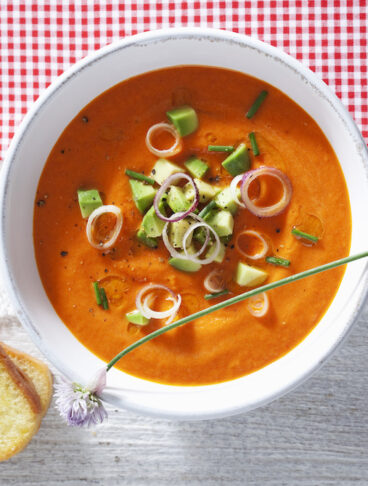 ciotola di gazpacho con crostino di pane su ripiano bianco con tovaglia a quadretti bianchi e rossi
