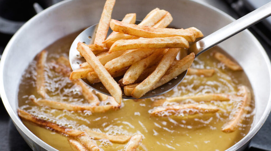 Una schiumarola di metallo solleva un gruppo di patatine fritte dorate e croccanti da una padella piena di olio bollente.