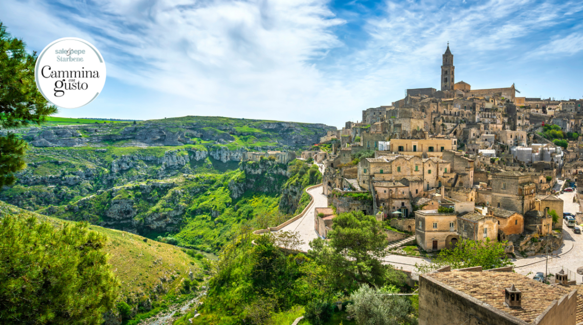 Veduta panoramica di Matera con i Sassi arroccati sulla destra e il profondo canyon verde della Gravina sulla sinistra, sotto un cielo azzurro con nuvole velate