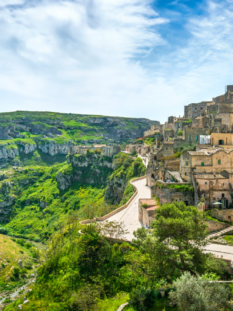 Veduta panoramica di Matera con i Sassi arroccati sulla destra e il profondo canyon verde della Gravina sulla sinistra, sotto un cielo azzurro con nuvole velate