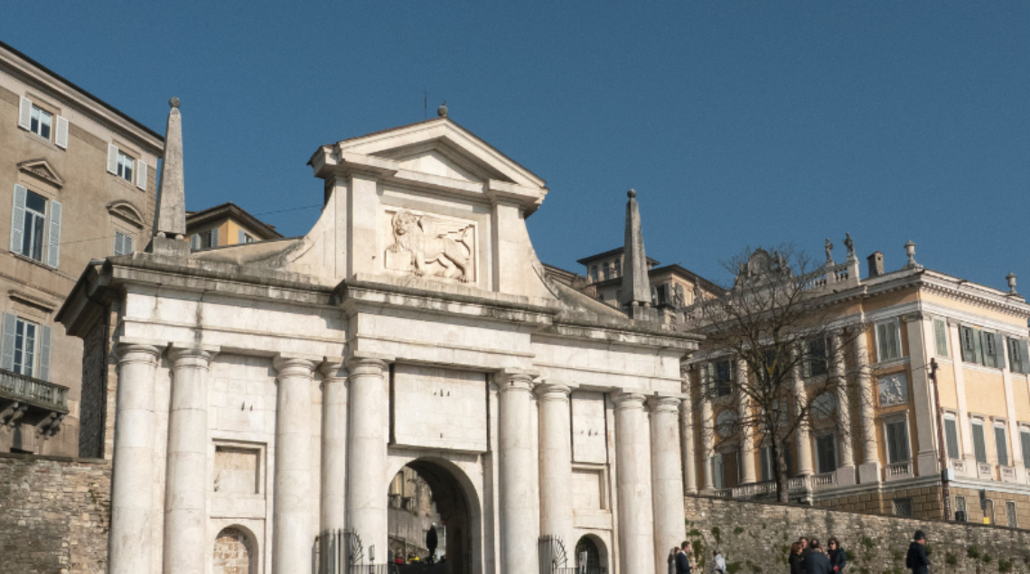 Vista prospettica della Porta San Giacomo in Città Alta a Bergamo sotto un cielo azzurro, con il Leone di San Marco in rilievo.