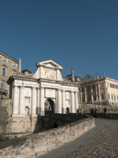 Vista prospettica della Porta San Giacomo in Città Alta a Bergamo sotto un cielo azzurro, con il Leone di San Marco in rilievo.