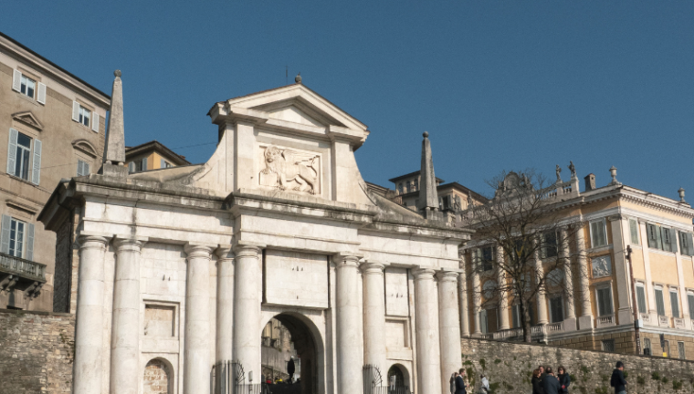 Vista prospettica della Porta San Giacomo in Città Alta a Bergamo sotto un cielo azzurro, con il Leone di San Marco in rilievo.
