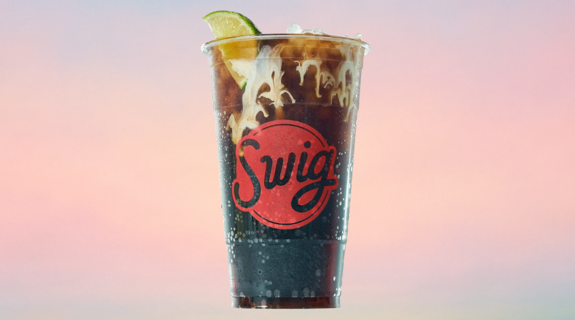 Bicchiere da asporto del brand Swig contenente una "dirty soda" scura con ghiaccio, una fetta di lime e una scenografica colata di crema di cocco che si mescola alla bevanda.