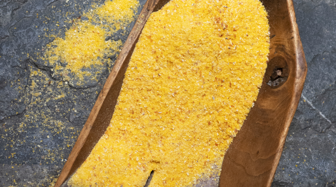 Farina di mais giallo a grana grossa versata in un contenitore di legno rustico su fondo in pietra scura. 