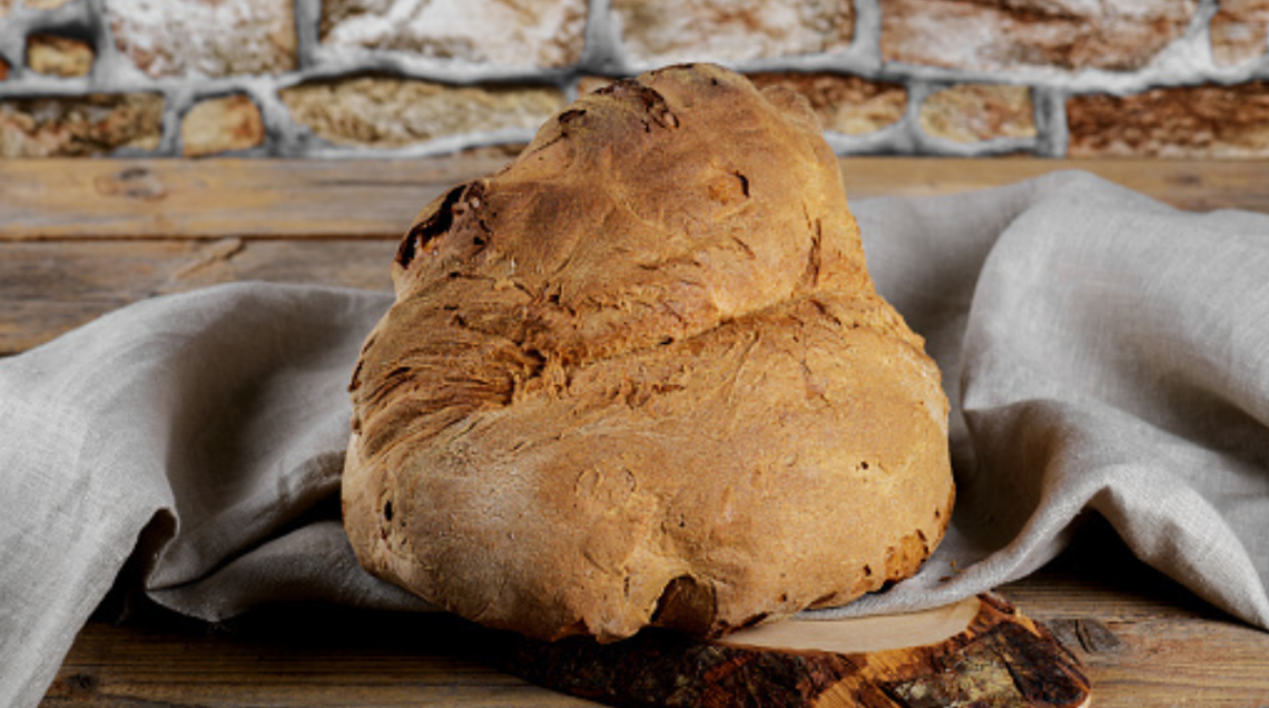 Una grande pagnotta di pane di Matera dalla forma tipica "a cornetto", con crosta bruna e croccante, appoggiata su un canovaccio grigio