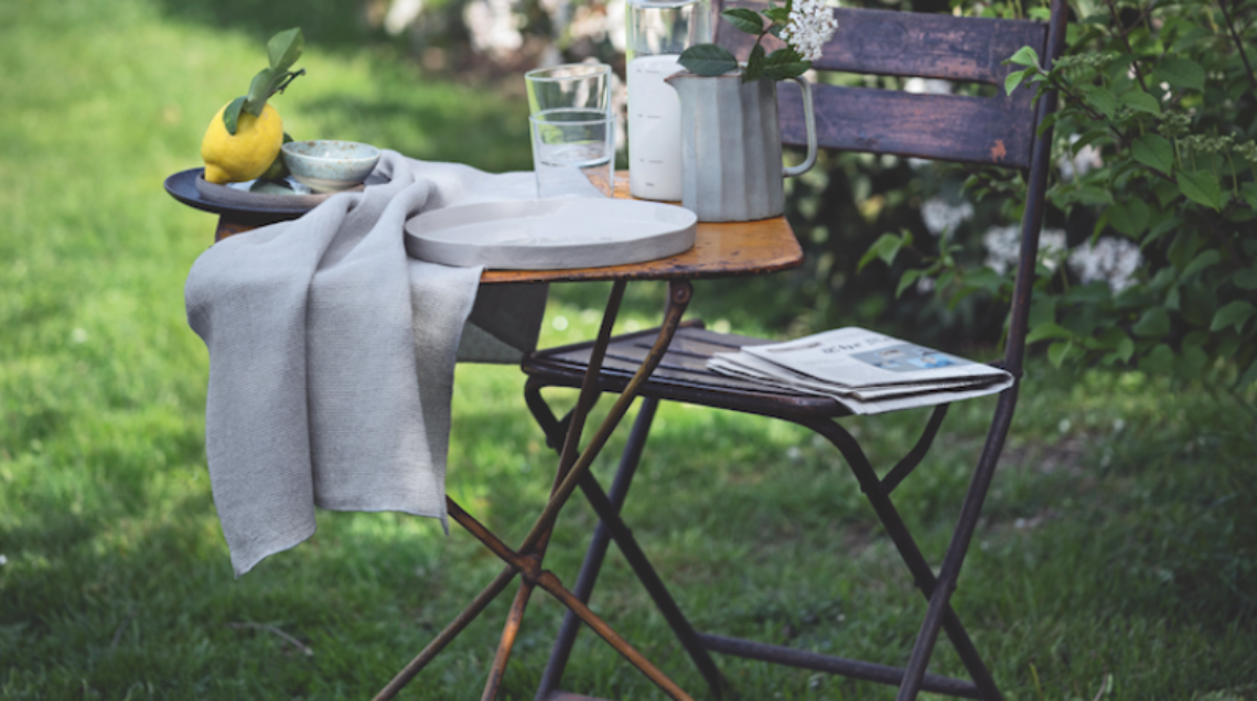 Un set da bistrot vintage in metallo arrugginito posizionato su un prato verde, apparecchiato con stoviglie e un limone, con un grande cespuglio di fiori bianchi sullo sfondo.