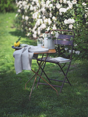 Un set da bistrot vintage in metallo arrugginito posizionato su un prato verde, apparecchiato con stoviglie e un limone, con un grande cespuglio di fiori bianchi sullo sfondo.