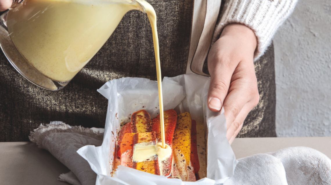 Una persona versa una crema gialla e densa sopra strati di carote colorate disposte in uno stampo da plumcake foderato con carta forno. In primo piano, una piccola ciotola di spezie macinate.