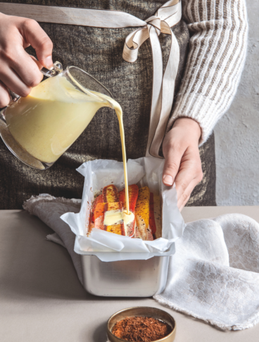 Una persona versa una crema gialla e densa sopra strati di carote colorate disposte in uno stampo da plumcake foderato con carta forno. In primo piano, una piccola ciotola di spezie macinate.