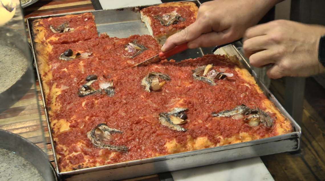 Mano che taglia un trancio di sardenaira, che mostra la struttura alta, la mollica bianca e il condimento ricco in superficie