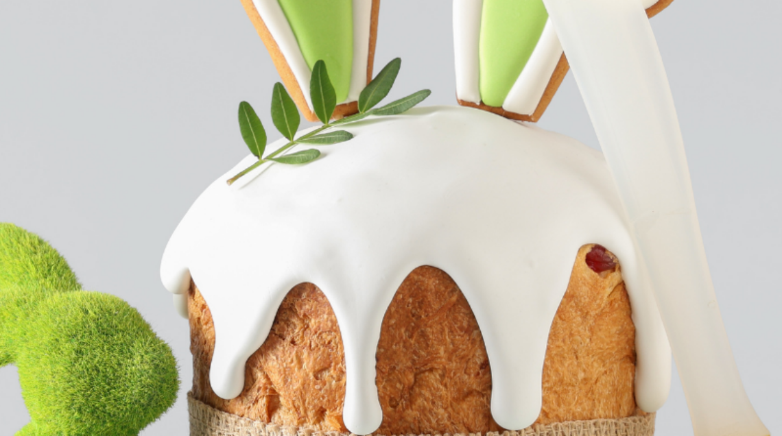 Dolce pasquale decorato con glassa bianca e biscotti a forma di orecchie di coniglio, circondato da uova colorate e un coniglietto di erba sintetica su sfondo grigio