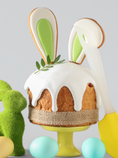 Dolce pasquale decorato con glassa bianca e biscotti a forma di orecchie di coniglio, circondato da uova colorate e un coniglietto di erba sintetica su sfondo grigio