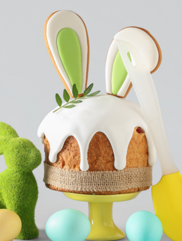 Dolce pasquale decorato con glassa bianca e biscotti a forma di orecchie di coniglio, circondato da uova colorate e un coniglietto di erba sintetica su sfondo grigio