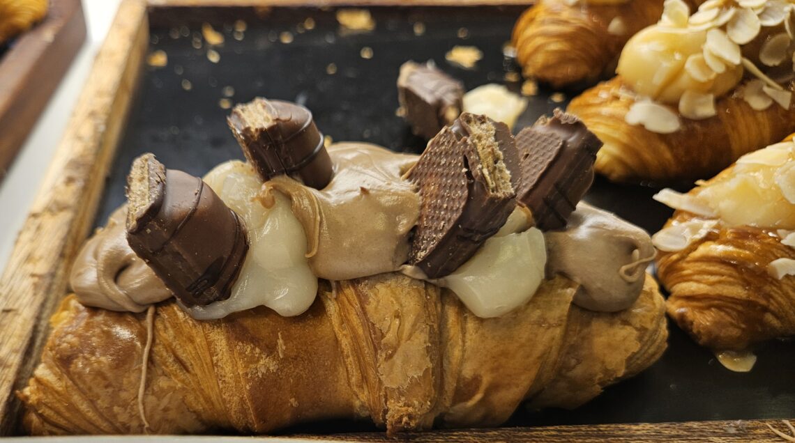 Croissant artigianale farcito con crema bicolore (latte e nocciola) e decorato con pezzi di cioccolato Kinder Bueno, esposto su un vassoio di legno.