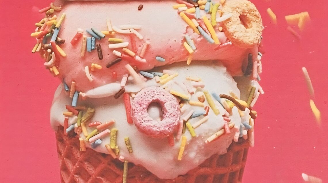 Primo piano di un cono gelato a tre gusti (menta, fragola e vaniglia) decorato con piccoli donut colorati, zuccherini multicolore e lenti di cioccolato su sfondo rosso vivace.