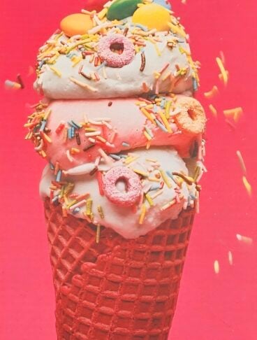 Primo piano di un cono gelato a tre gusti (menta, fragola e vaniglia) decorato con piccoli donut colorati, zuccherini multicolore e lenti di cioccolato su sfondo rosso vivace.