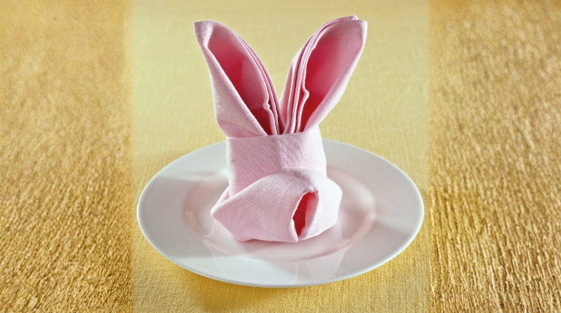 Tovagliolo di stoffa rosa piegato artisticamente a forma di coniglio con lunghe orecchie, appoggiato su un piatto bianco
