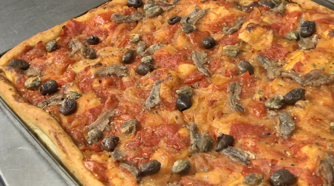 Macro sulla superficie della sardenaira cotta: dettaglio di olive taggiasche lucide, acciughe e una pioggia di origano secco.