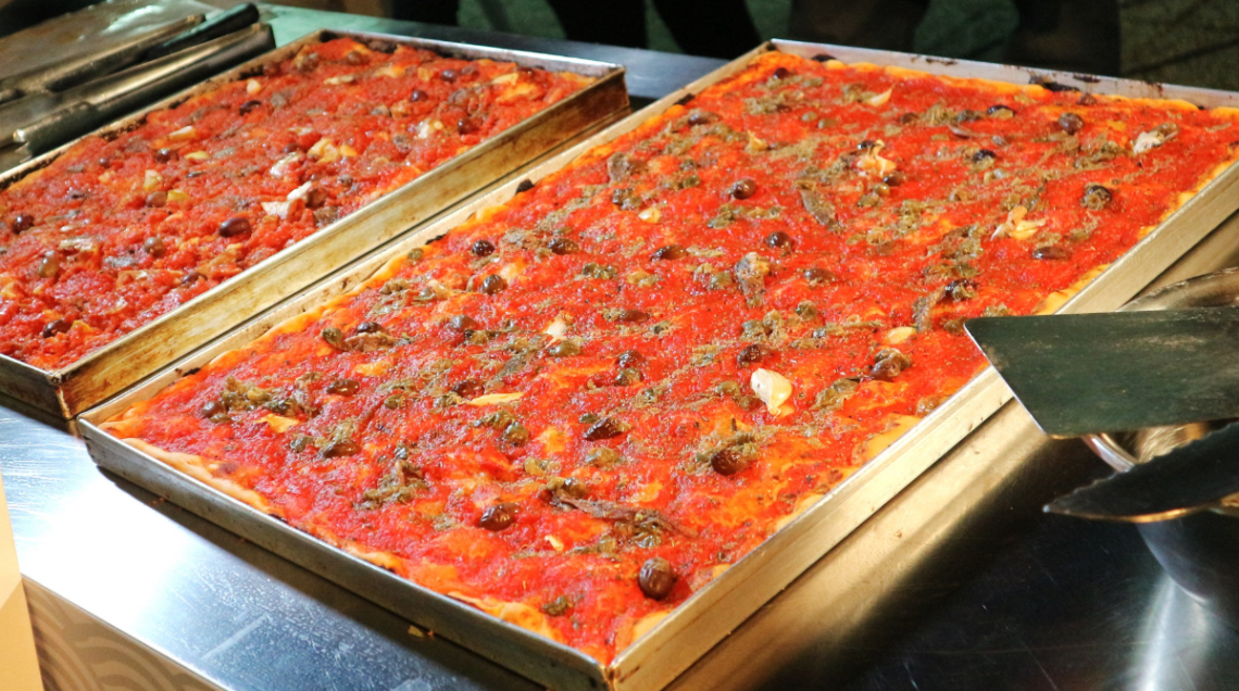 Una teglia rettangolare colma di sardenaira appena sfornata, guarnita con olive, aglio, acciughe e origano