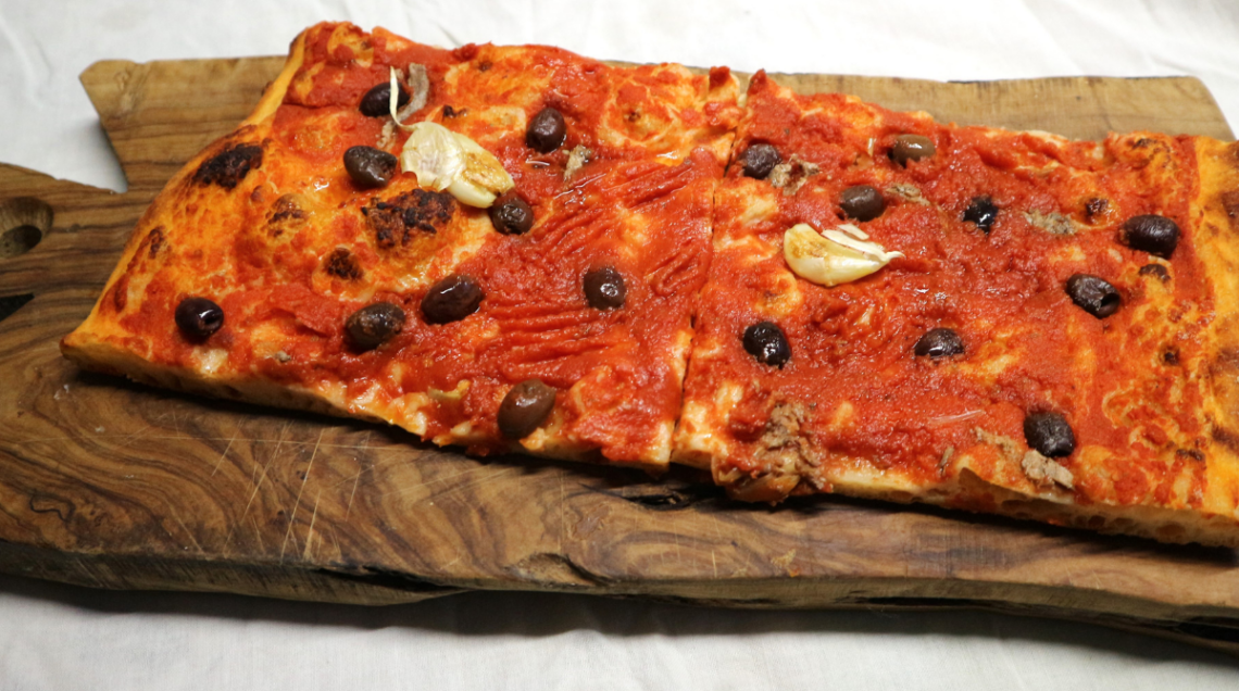 Tranci di Sardenaira alla sanremasca disposti su un tagliere di legno rustico, guarniti con olive taggiasche e origano