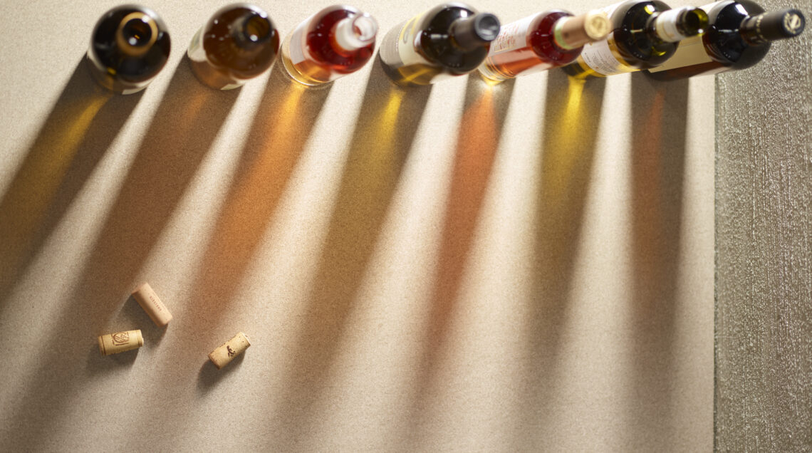 Una serie di bottiglie di vino allineate su una superficie testurizzata, proiettando lunghe ombre proiettate verso destra. Sono visibili tre tappi di sughero in basso a destra