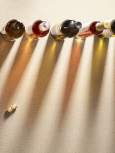 Una serie di bottiglie di vino allineate su una superficie testurizzata, proiettando lunghe ombre proiettate verso destra. Sono visibili tre tappi di sughero in basso a destra