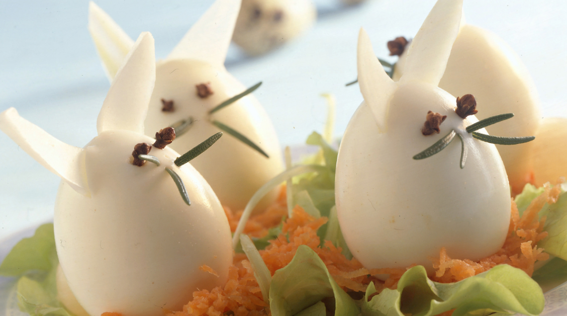 Quattro uova sode decorate come coniglietti con orecchie di sedano e baffi di rosmarino su un letto di carote