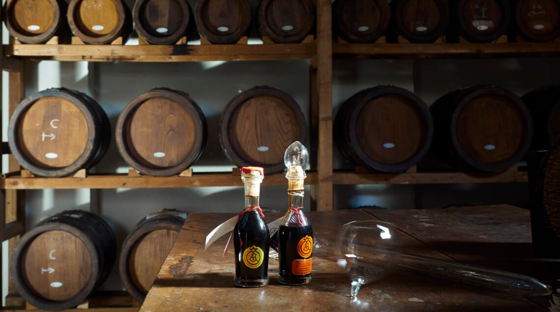 Piccole botti di legno antico per l'Aceto Balsamico Tradizionale disposte su scaffali, con due bottiglie tipiche in primo piano.