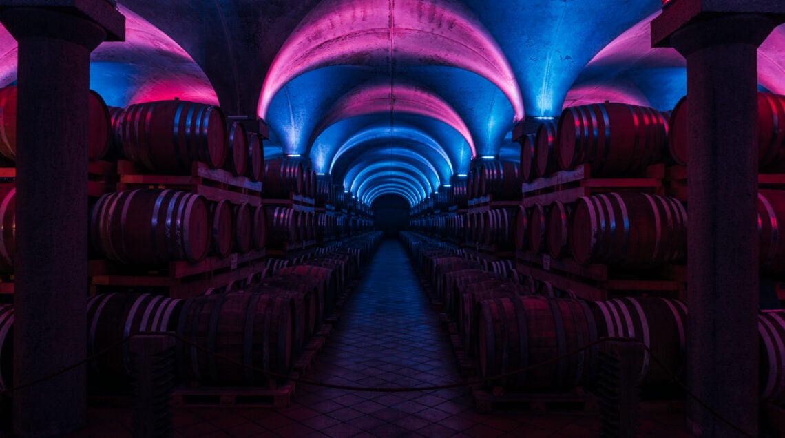 Cantina sotterranea con volte a crociera illuminate da luci soffuse blu e viola, file di botti di legno per l'invecchiamento della grappa. 