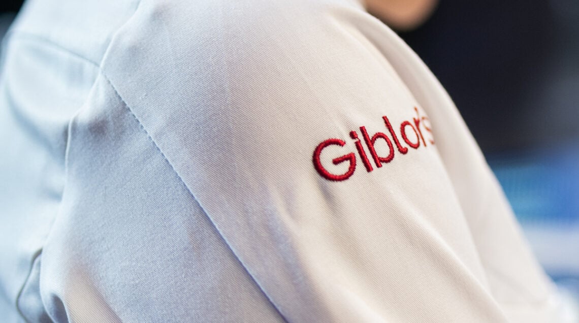 Inquadratura laterale di una giacca da cuoco bianca con il logo "Giblor's" ricamato in rosso sulla spalla