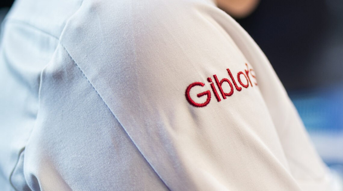 Inquadratura laterale di una giacca da cuoco bianca con il logo "Giblor's" ricamato in rosso sulla spalla