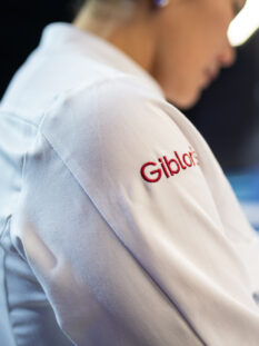 Inquadratura laterale di una giacca da cuoco bianca con il logo "Giblor's" ricamato in rosso sulla spalla
