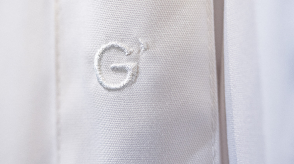 Primo piano di un ricamo bianco a forma di lettera "G" stilizzata su tessuto bianco di una divisa professionale Giblor’s.