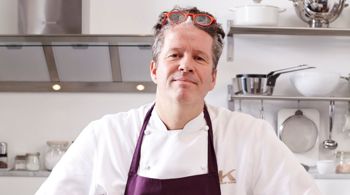 Ernst Knam in cucina con grembiule bordeaux e occhiali rossi.