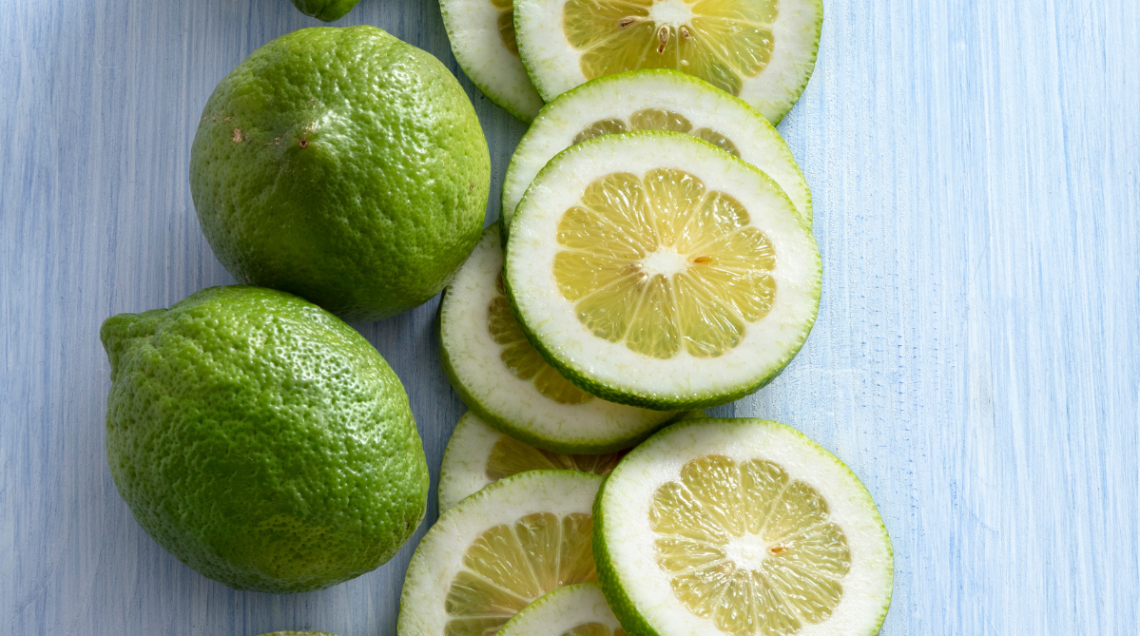 Limoni verdelli interi e tagliati a fette circolari su un tavolo di legno azzurro, che mostrano la buccia verde e la polpa chiara.