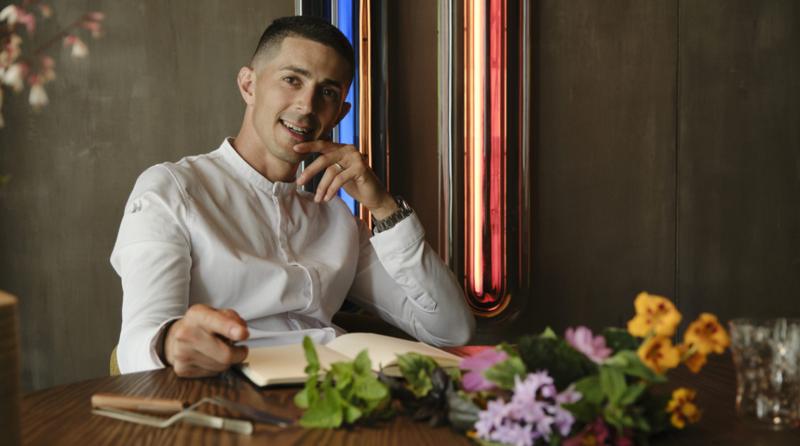 Lo chef Michelangelo Mammoliti seduto a un tavolo con un taccuino e fiori colorati.