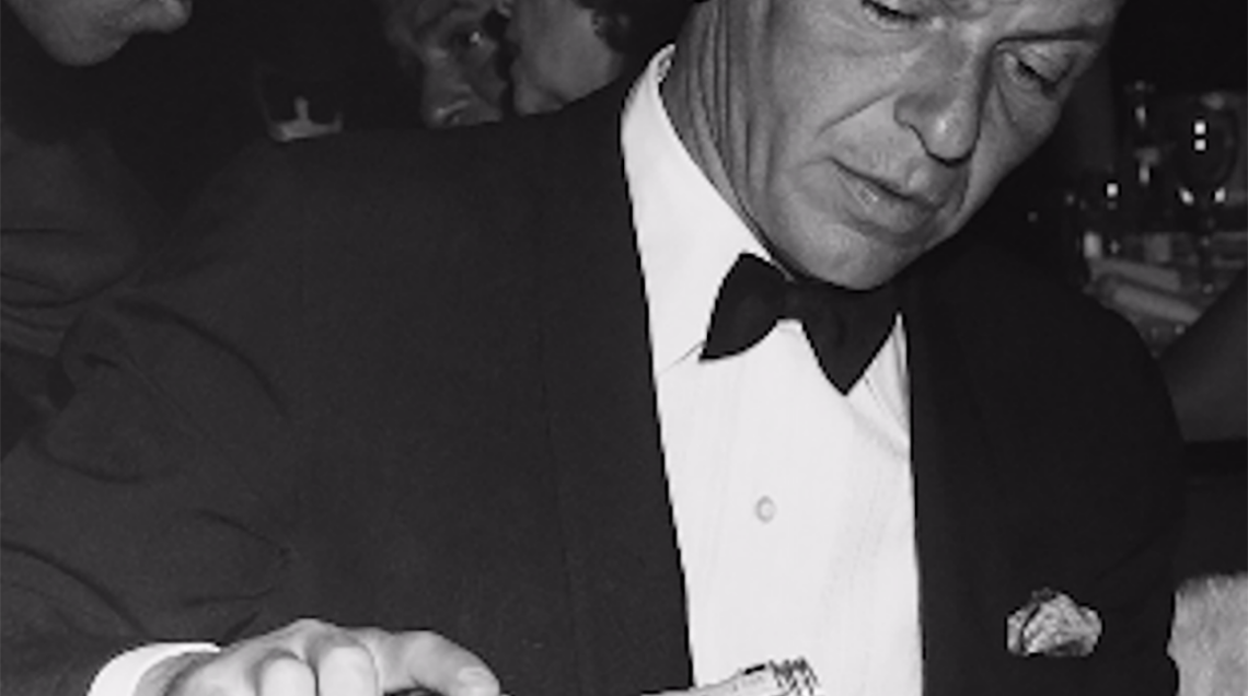 Sinatra in smoking. seduto a tavola. si versa un Jack Daniel's