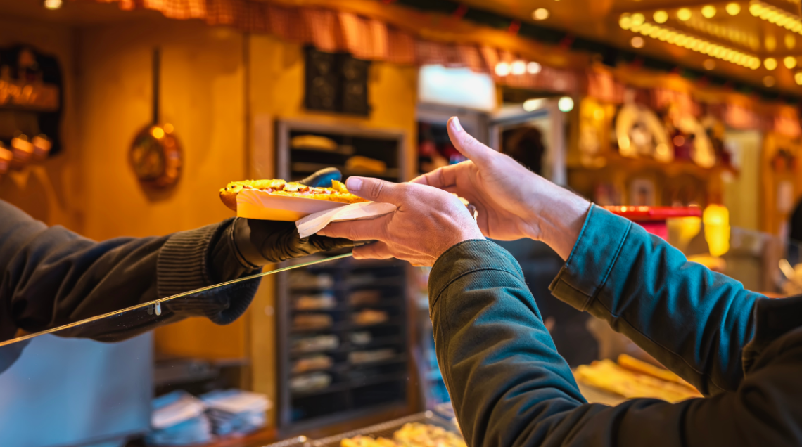 Inquadratura ravvicinata di un cliente che prende un panino farcito da un venditore attraverso lo sportello in vetro di un chiosco in legno illuminato.