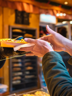 Inquadratura ravvicinata di un cliente che prende un panino farcito da un venditore attraverso lo sportello in vetro di un chiosco in legno illuminato.