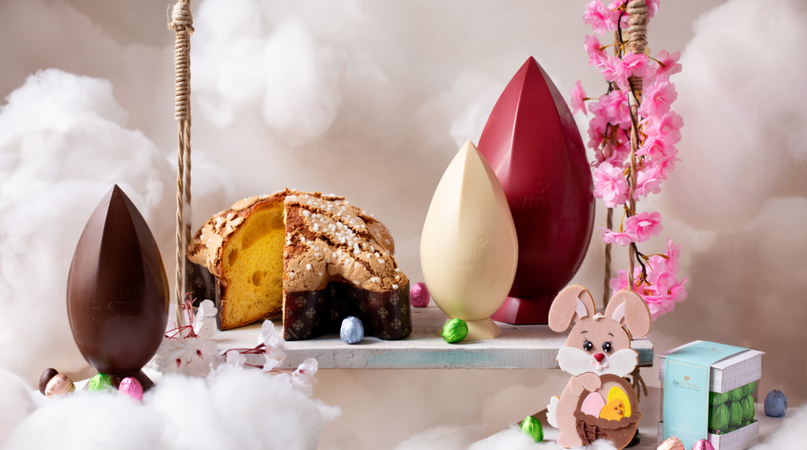 Composizione di uova di cioccolato di diversi colori e una colomba artigianale Da Vittorio su un'altalena circondata da nuvole bianche e fiori rosa.