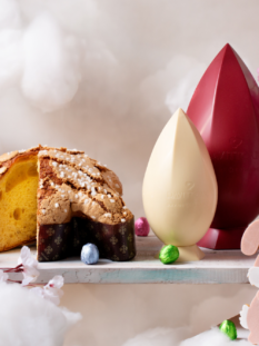 Composizione di uova di cioccolato di diversi colori e una colomba artigianale Da Vittorio su un'altalena circondata da nuvole bianche e fiori rosa.