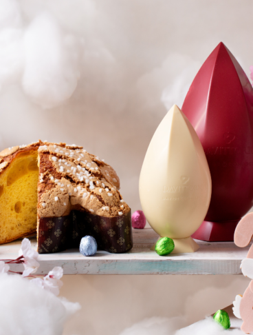 Composizione di uova di cioccolato di diversi colori e una colomba artigianale Da Vittorio su un'altalena circondata da nuvole bianche e fiori rosa.