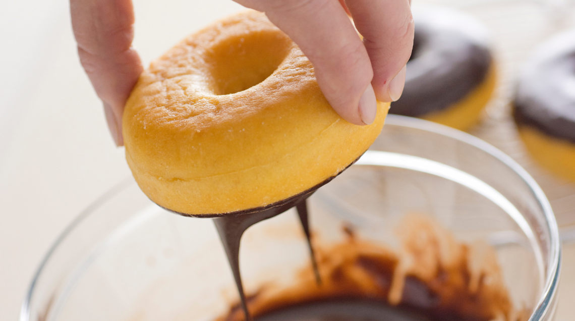 Una mano che immerge una ciambella dorata in una ciotola di cioccolato fondente fuso, con altre ciambelle già pronte sullo sfondo.